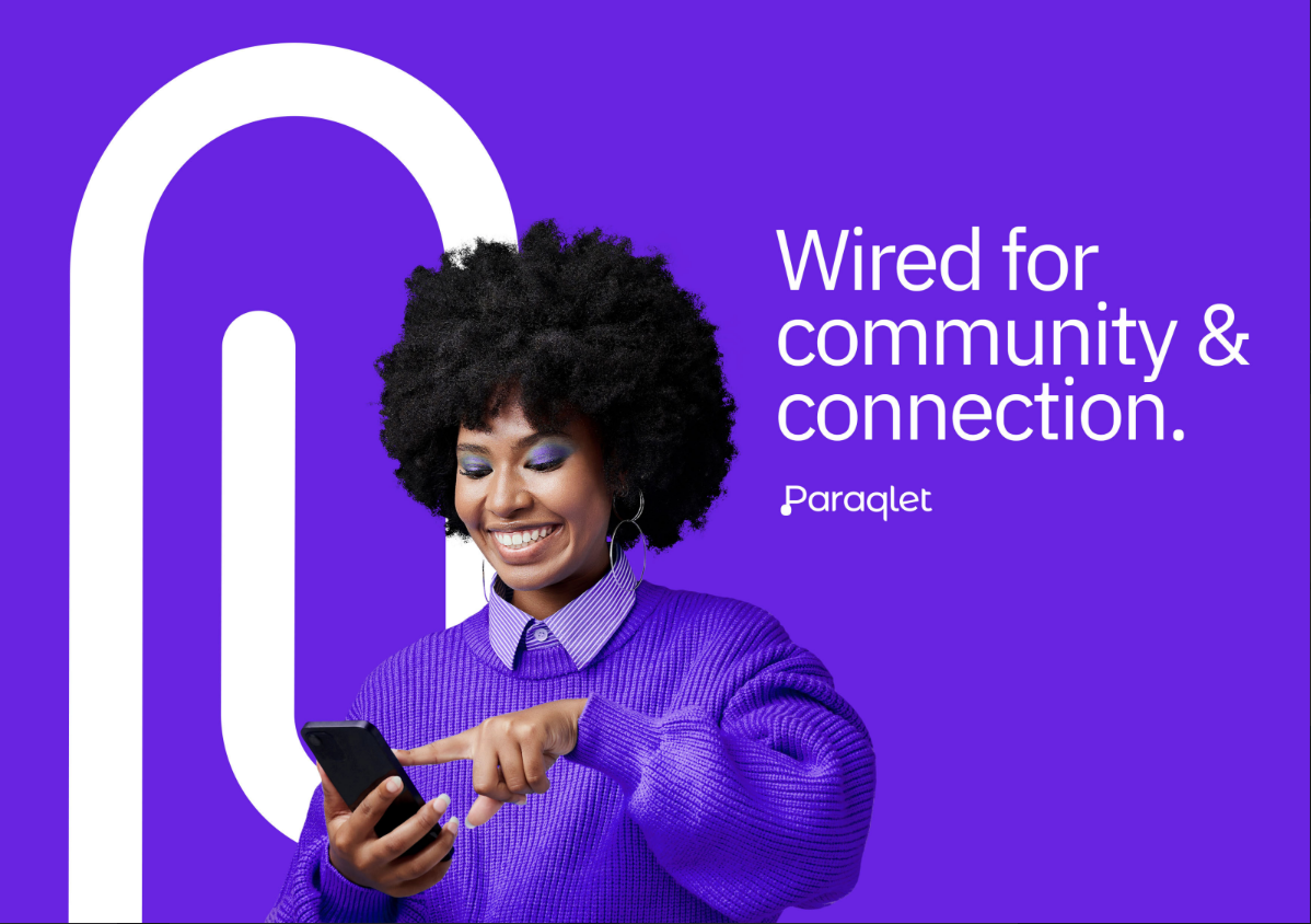 Wired for community & connection. Paraqlet. A Black woman wearing a purple sweater and lilac eyeshadow smiles as she taps her phone.